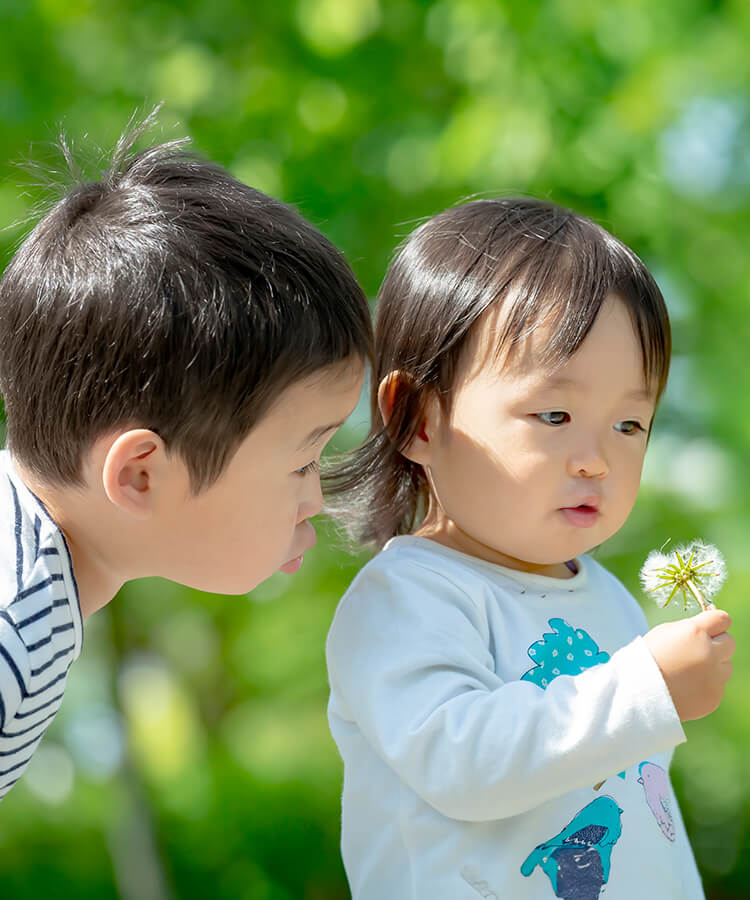 二人の子供が緑の中で楽しそうにタンポポの綿毛を吹いている写真